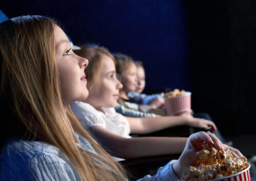 View from side of beautiful little girl sitting in cinema and watching interesting film. Young spectator resting with friends and eating popcorn. Concept of having fun and entertainment.