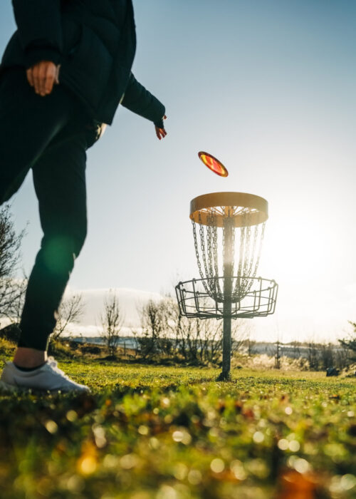 Low angle shot of a woman tossing a disc into the basket goal. Female playing disc golf game outdoors in a park.